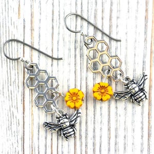 May include: Silver-coloured bee-themed earrings. Each earring features a honeycomb charm, a yellow flower bead, and a detailed bee pendant. The earrings hang from silver-coloured ear wires, creating a charming and nature-inspired accessory.
