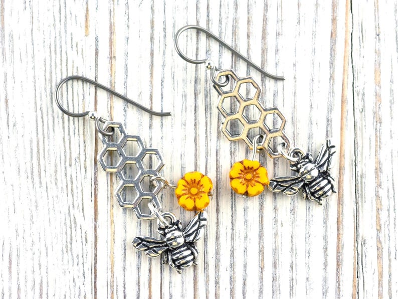 May include: Silver-coloured bee-themed earrings. Each earring features a honeycomb charm, a yellow flower bead, and a detailed bee pendant. The earrings hang from silver-coloured ear wires, creating a charming and nature-inspired accessory.