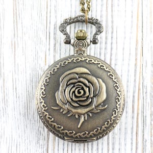 May include: Antique bronze-colored pocket watch with a detailed rose design on the face. The watch has a decorative border and is suspended from a matching chain. A classic, vintage-style accessory.