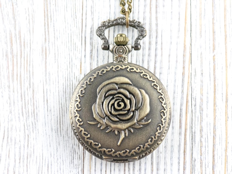 May include: Antique bronze-colored pocket watch with a detailed rose design on the face. The watch has a decorative border and is suspended from a matching chain. A classic, vintage-style accessory.