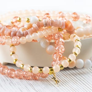 May include: A set of pink and gold beaded bracelets with a star charm. The bracelets are made of different types of beads, including sunstone, glass, and gold.