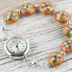 May include: A silver pocket watch with a white face and black hands attached to a chain with orange and green enamel beads. The watch face reads "GENEVA QUARTZ".