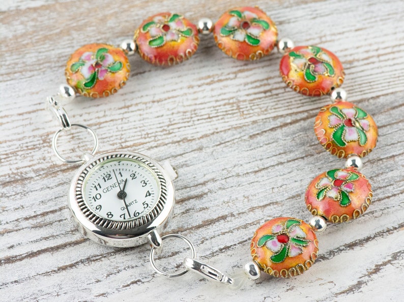 May include: A silver pocket watch with a white face and black hands attached to a chain with orange and green enamel beads. The watch face reads "GENEVA QUARTZ".