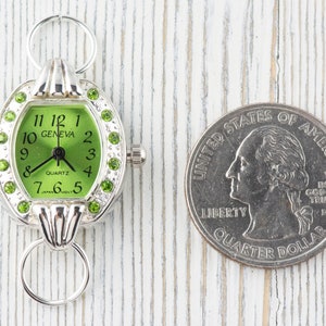 Op de afbeelding: Een zilveren horloge met een groene wijzerplaat en strass-accenten. Het horloge is ongeveer zo groot als een Amerikaanse kwart.