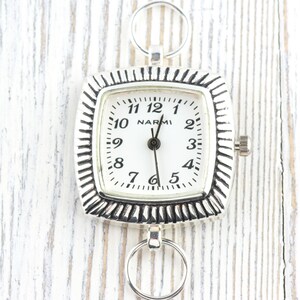 May include: A square silver pocket watch with a white face and black numerals. The watch has a textured bezel and two circular rings for attachment. The brand name "NARMI" is printed on the watch face.