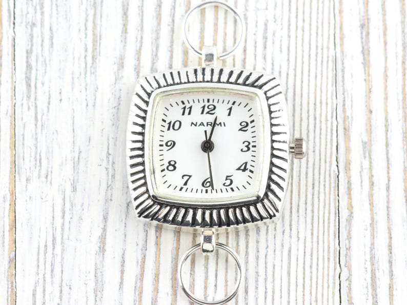 May include: A square silver pocket watch with a white face and black numerals. The watch has a textured bezel and two circular rings for attachment. The brand name "NARMI" is printed on the watch face.