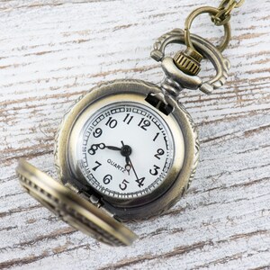 May include: A vintage-style pocket watch with a brass finish and a white face. The watch has Roman numeral hour markers and a black second hand. The watch is open and the chain is visible.