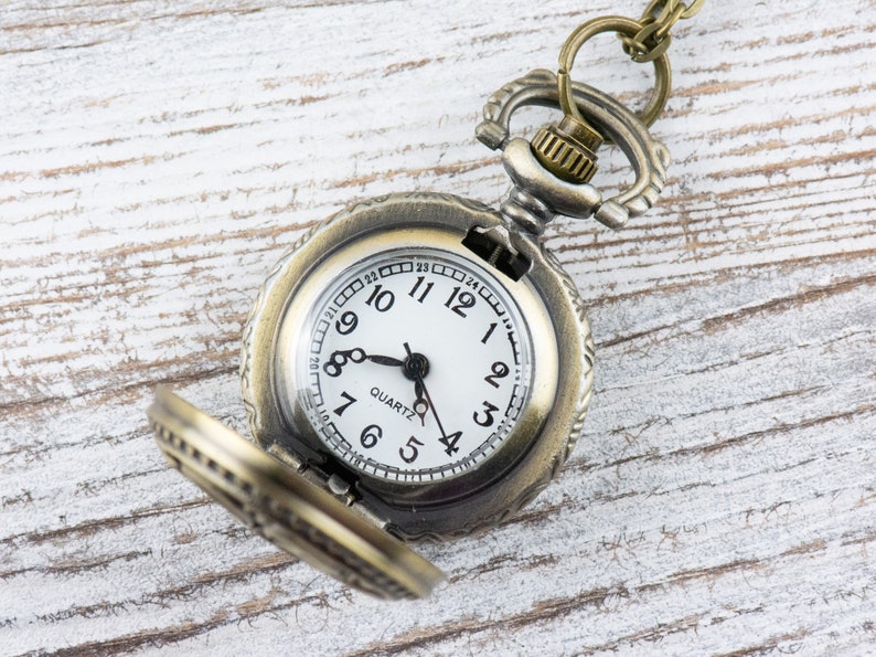 May include: A vintage-style pocket watch with a brass finish and a white face. The watch has Roman numeral hour markers and a black second hand. The watch is open and the chain is visible.