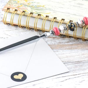 May include: A decorative letter opener with a silver blade and a beaded handle. The handle features pink, black, and silver beads. The letter opener rests on a white envelope with a gold heart sticker, next to a floral notebook.