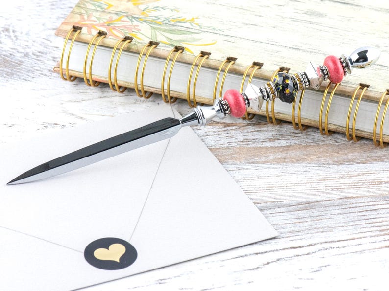 May include: A decorative letter opener with a silver blade and a beaded handle. The handle features pink, black, and silver beads. The letter opener rests on a white envelope with a gold heart sticker, next to a floral notebook.