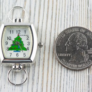 May include: Silver-toned pocket watch with a Christmas tree design on the face, next to a quarter. The watch has a rectangular face with a curved shape and a ring at the top and bottom. The watch face displays the time and the brand name "NARMI".