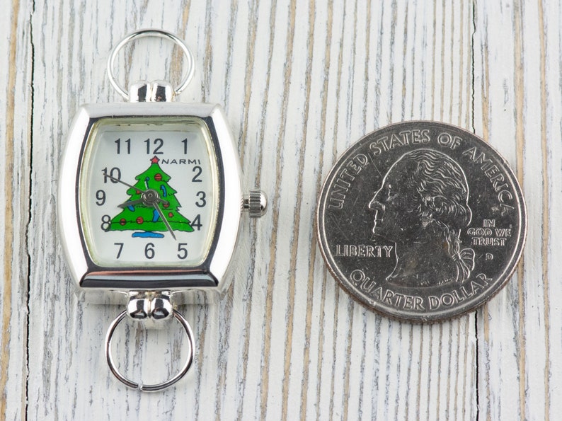 May include: Silver-toned pocket watch with a Christmas tree design on the face, next to a quarter. The watch has a rectangular face with a curved shape and a ring at the top and bottom. The watch face displays the time and the brand name "NARMI".