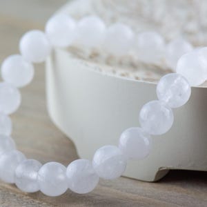May include: A white beaded bracelet made of round, opaque, white stones. The bracelet is lying on a white surface.