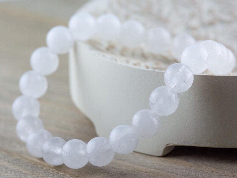 May include: A white beaded bracelet made of round, opaque, white stones. The bracelet is lying on a white surface.