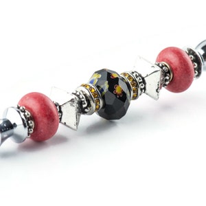 May include: A decorative bottle stopper with a silver stem and a series of beads. The beads include red, silver, and a black faceted bead with yellow and floral accents. The stopper has a silver, rounded top.
