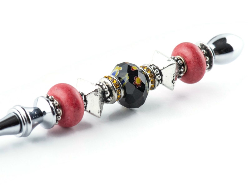 May include: A decorative bottle stopper with a silver stem and a series of beads. The beads include red, silver, and a black faceted bead with yellow and floral accents. The stopper has a silver, rounded top.