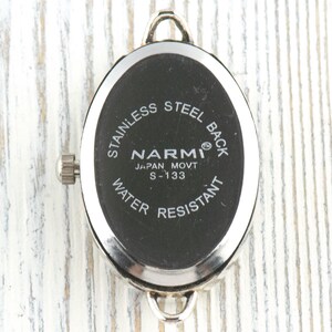 May include: A close-up of the back of a watch with a black oval face. The watch has a silver metal frame and a silver crown. The text on the watch face reads "STAINLESS STEEL BACK", "NARMI JAPAN MOVT S-133", and "WATER RESISTANT".