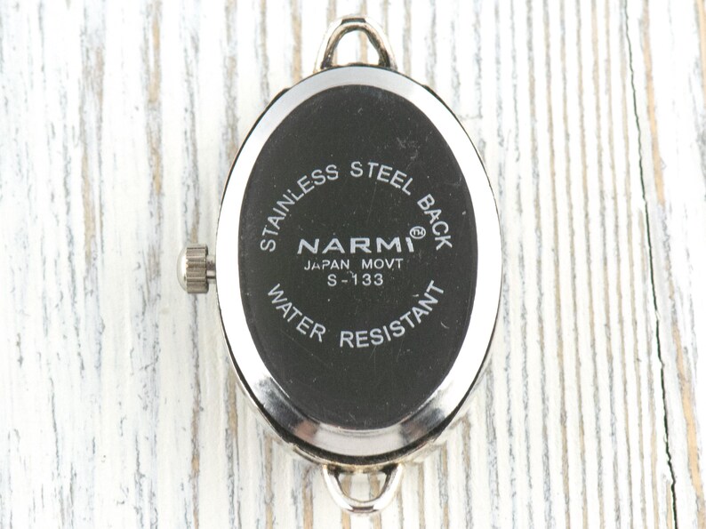 May include: A close-up of the back of a watch with a black oval face. The watch has a silver metal frame and a silver crown. The text on the watch face reads "STAINLESS STEEL BACK", "NARMI JAPAN MOVT S-133", and "WATER RESISTANT".
