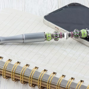 K&ouml;nnte beinhalten: Ein silberner Stift mit einem gr&uuml;nen, rosa und schwarzen Perlenmuster. Der Stift hat eine Stiftspitze und liegt auf einem Notizbuch mit goldener Spiralbindung.