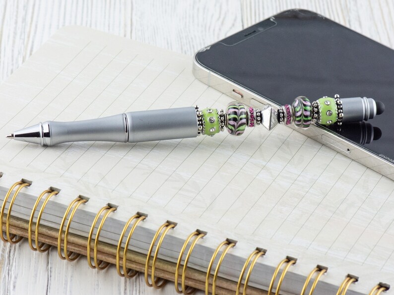 K&ouml;nnte beinhalten: Ein silberner Stift mit einem gr&uuml;nen, rosa und schwarzen Perlenmuster. Der Stift hat eine Stiftspitze und liegt auf einem Notizbuch mit goldener Spiralbindung.