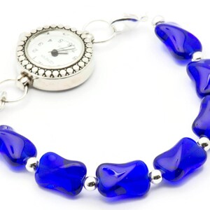 Pode incluir: Um rel&oacute;gio de pulso prateado com um mostrador branco e ponteiros pretos presos a uma pulseira feita de contas de vidro azul.