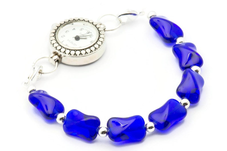 Pode incluir: Um rel&oacute;gio de pulso prateado com um mostrador branco e ponteiros pretos presos a uma pulseira feita de contas de vidro azul.