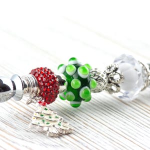 May include: A decorative bottle stopper with a silver base and a series of colorful beads. The beads include a red, jeweled bead, a green and white polka-dot bead, and a clear, faceted bead. A small Christmas tree charm hangs from the red bead.