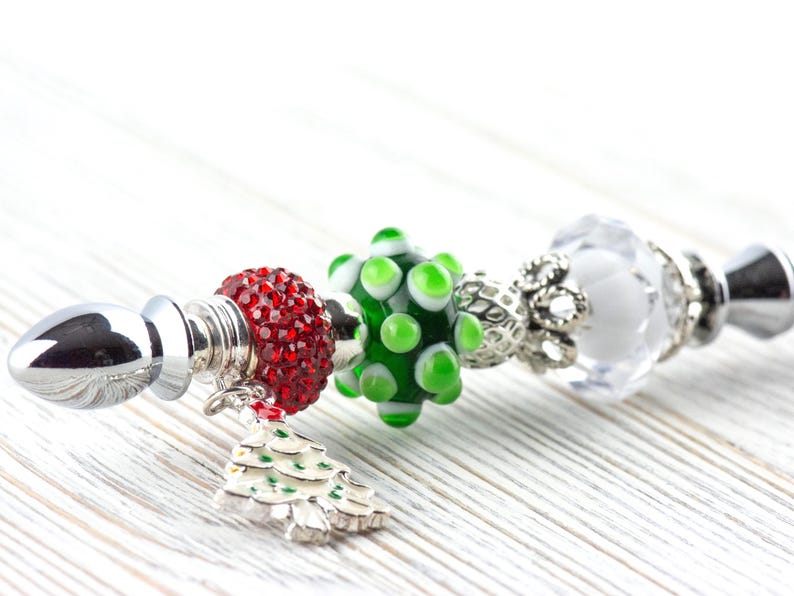May include: A decorative bottle stopper with a silver base and a series of colorful beads. The beads include a red, jeweled bead, a green and white polka-dot bead, and a clear, faceted bead. A small Christmas tree charm hangs from the red bead.