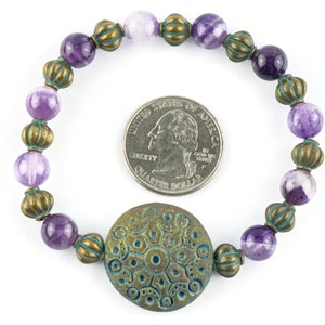 May include: Purple and white agate beads with bronze accents and a large bronze charm with a blue and green design. The bracelet is about the size of a US quarter.
