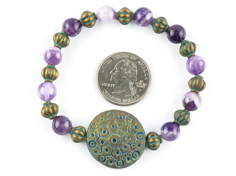 May include: Purple and white agate beads with bronze accents and a large bronze charm with a blue and green design. The bracelet is about the size of a US quarter.