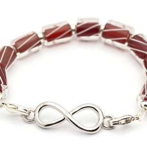 K&ouml;nnte beinhalten: Ein Armband aus roten und wei&szlig;en, gestreiften Glasperlen mit einem Unendlichkeits-Anh&auml;nger. Das Armband ist aus Silber und hat einen Karabinerverschluss.