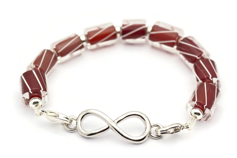 K&ouml;nnte beinhalten: Ein Armband aus roten und wei&szlig;en, gestreiften Glasperlen mit einem Unendlichkeits-Anh&auml;nger. Das Armband ist aus Silber und hat einen Karabinerverschluss.