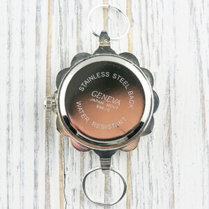 May include: The back of a silver watch with a round face and a scalloped edge. The watch face has the text "Stainless Steel Back Geneva Japan Movt BSR-3 Water Resistant" printed on it.