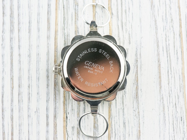 May include: The back of a silver watch with a round face and a scalloped edge. The watch face has the text "Stainless Steel Back Geneva Japan Movt BSR-3 Water Resistant" printed on it.