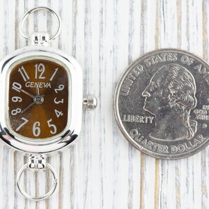 May include: A silver Geneva watch with a brown face and white numerals, next to a U.S. quarter. The watch has a rectangular face with rounded corners and a small crown. The quarter shows a profile of George Washington.