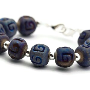 Pu&ograve; includere: Un braccialetto blu e argento con una serie di perline in ceramica blu scuro. Ogni perlina ha un motivo a spirale intagliato.