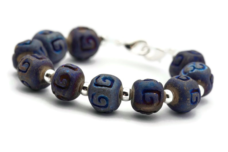 Pu&ograve; includere: Un braccialetto blu e argento con una serie di perline in ceramica blu scuro. Ogni perlina ha un motivo a spirale intagliato.