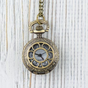 Puede incluir: Un reloj de bolsillo dorado con una esfera blanca y agujas negras. El reloj tiene un dise&ntilde;o decorativo y ornamentado alrededor del borde de la esfera y est&aacute; colgado de una cadena.