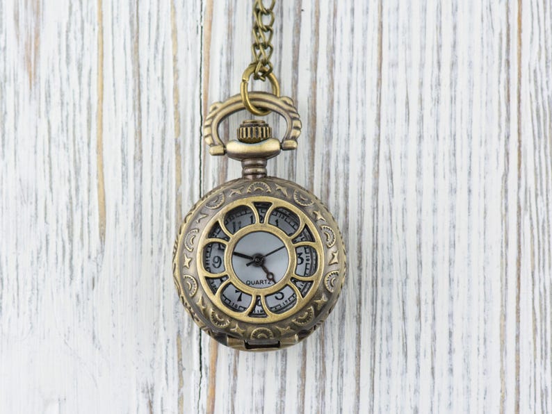Puede incluir: Un reloj de bolsillo dorado con una esfera blanca y agujas negras. El reloj tiene un dise&ntilde;o decorativo y ornamentado alrededor del borde de la esfera y est&aacute; colgado de una cadena.