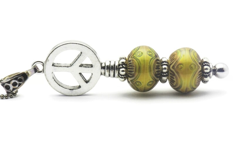 Puede incluir: Un colgante de plata con forma de signo de paz con una cadena y dos cuentas de vidrio verde y marr&oacute;n con un separador de plata entre ellas.