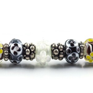 May include: A close-up of a bracelet with a variety of colorful glass beads, including black and white patterned beads, yellow and brown beads, and white beads with a floral design.