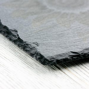 K&ouml;nnte beinhalten: Nahaufnahme einer schwarzen Schiefer-Servierplatte mit einer rauen, strukturierten Oberfl&auml;che. Die Platte ist schr&auml;g und teilweise sichtbar vor einem wei&szlig;en Holzhintergrund.