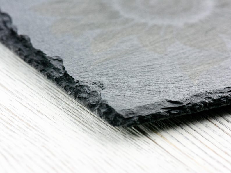 K&ouml;nnte beinhalten: Nahaufnahme einer schwarzen Schiefer-Servierplatte mit einer rauen, strukturierten Oberfl&auml;che. Die Platte ist schr&auml;g und teilweise sichtbar vor einem wei&szlig;en Holzhintergrund.