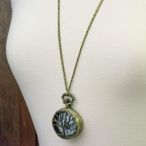May include: A bronze-coloured pocket watch pendant necklace. The watch face features a tree of life design. The chain is a delicate bronze colour. The watch is hanging on a white surface.