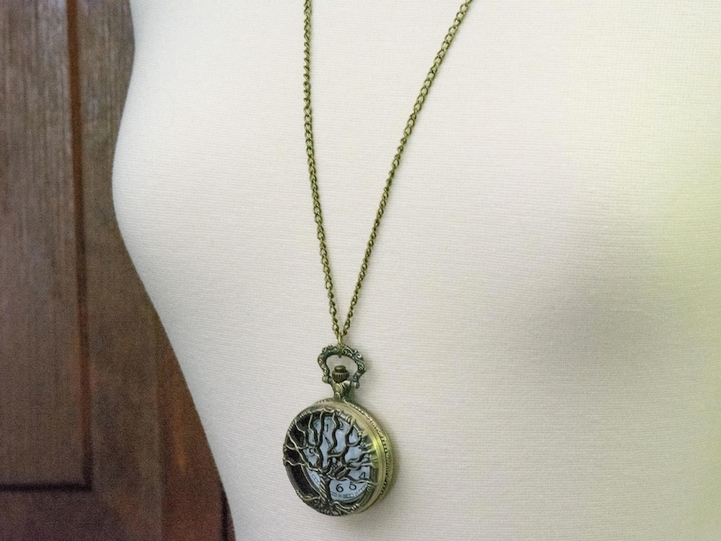 May include: A bronze-coloured pocket watch pendant necklace. The watch face features a tree of life design. The chain is a delicate bronze colour. The watch is hanging on a white surface.