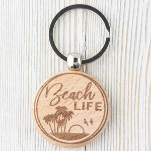 Pu&ograve; includere: Un portachiavi in legno con un anello portachiavi argentato. Il portachiavi &egrave; rotondo e ha le parole "Beach Life" incise su di esso. Ci sono palme e un tramonto incisi sul portachiavi.