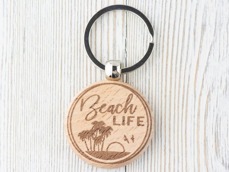 Pu&ograve; includere: Un portachiavi in legno con un anello portachiavi argentato. Il portachiavi &egrave; rotondo e ha le parole "Beach Life" incise su di esso. Ci sono palme e un tramonto incisi sul portachiavi.
