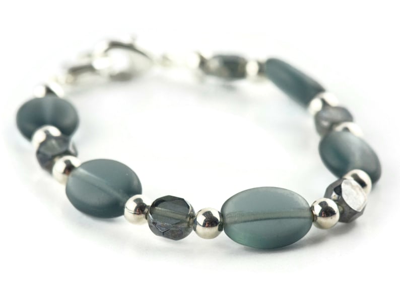 Pu&ograve; includere: Un braccialetto con perline ovali e sfaccettate grigio-verdi, alternate a piccole perline rotonde color argento. Il braccialetto &egrave; assicurato con una chiusura color argento. Le perline hanno un aspetto smerigliato.