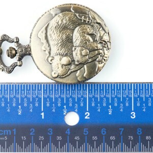 May include: A bronze pocket watch with a chain. The watch face features an engraved design of two bears hugging. The watch is measured against a ruler, showing it is approximately 2 inches in diameter.