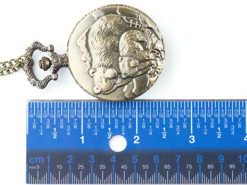 May include: A bronze pocket watch with a chain. The watch face features an engraved design of two bears hugging. The watch is measured against a ruler, showing it is approximately 2 inches in diameter.